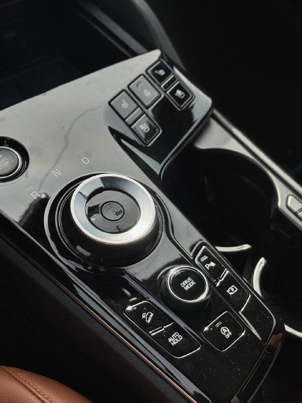 Селектор трансмиссии и центральная консоль KIA Sportage: роторный переключатель коробки передач, кнопки управления обогревом и медиасистемой в чёрной глянцевой отделке.