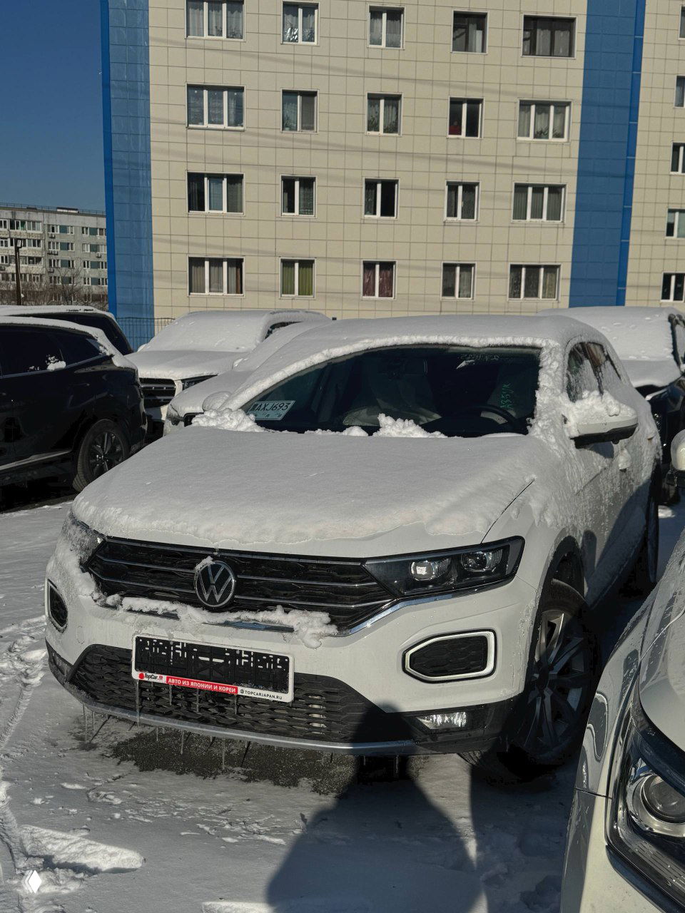 Белый Volkswagen T‑ROC, покрытый снегом, припаркован у жилого дома — вид спереди с номерной рамкой и наледью под бампером.