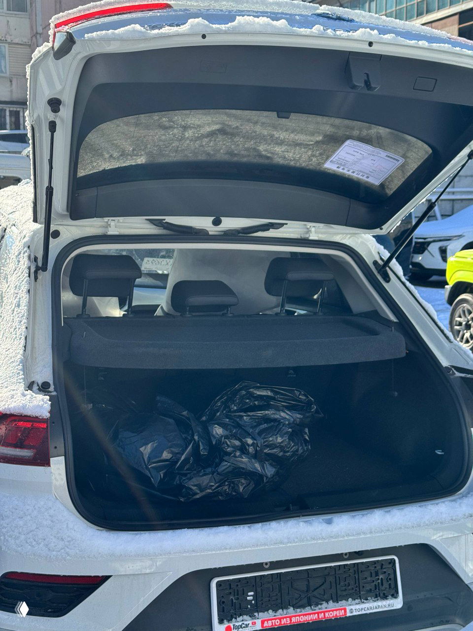 Открытый багажник белого Volkswagen T‑ROC с полкой и пакетами внутри, вид сзади машины на снежной стоянке после выгрузки.