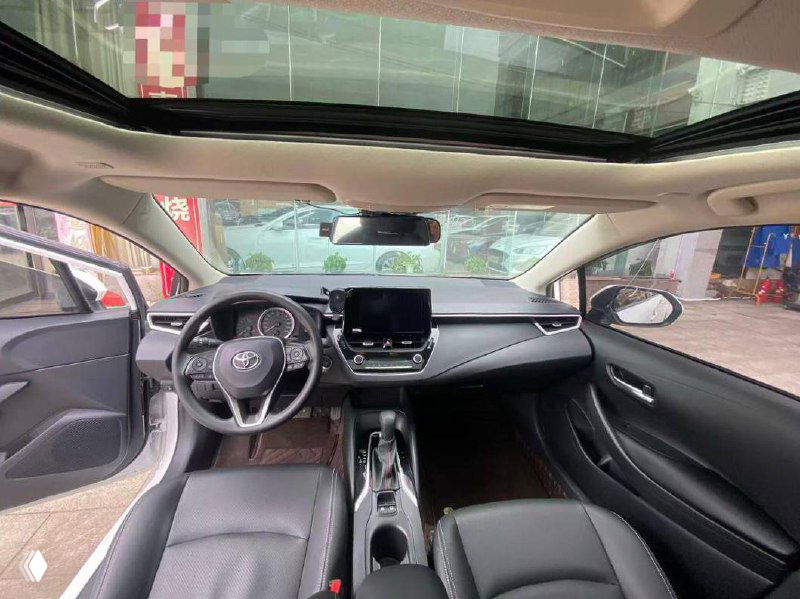 Интерьер Toyota Corolla 2023: общий план салона, передняя панель, мультимедиа и часть панорамной крыши; левый руль и кожаные сиденья.