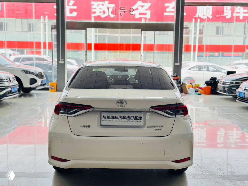 Фото задней части белой Toyota Corolla 2022 в шоуруме — вид на багажник, фон с витринами и соседними автомобилями внутри салона.