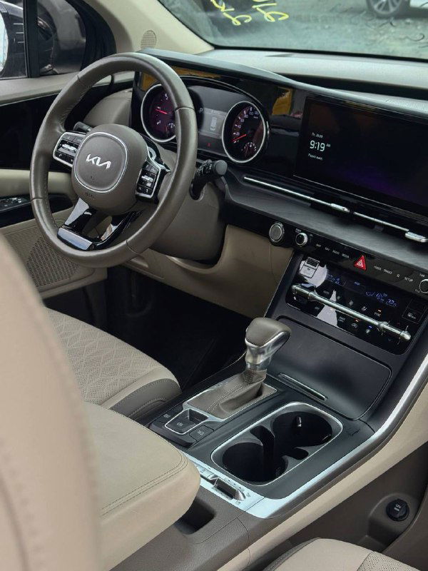 Передняя панель и центральная консоль KIA Carnival 2022: крупный план мультимедиа, селектор коробки и подстаканники в салоне автомобиля.