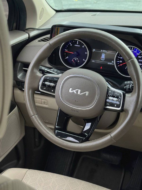 Руль с логотипом KIA и приборная панель KIA Carnival 2022, вид на спидометр и тахометр в салоне автомобиля с бежевой отделкой.