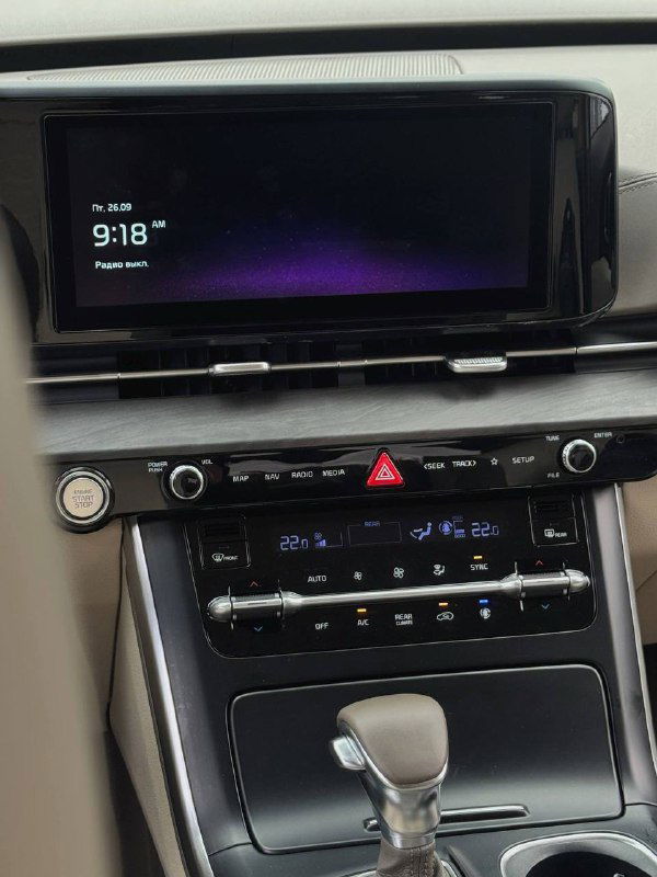 Широкий дисплей мультимедийной системы и панель управления климатом KIA Carnival, отображение времени и элементов интерфейса в салоне.