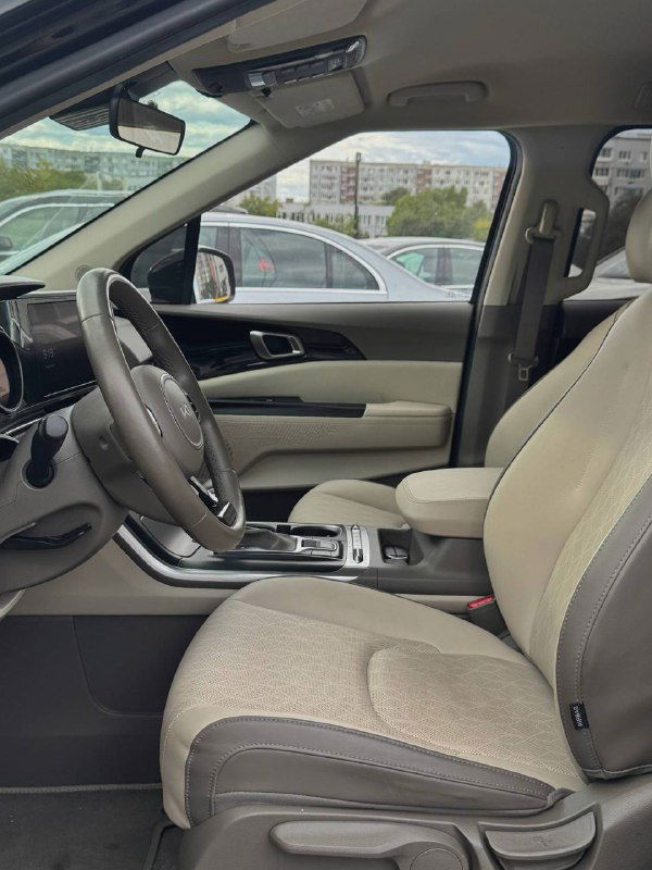Боковой вид на переднее сиденье и дверную панель KIA Carnival 2022 с бежевой обивкой, рукояткой и панелью управления стеклами.