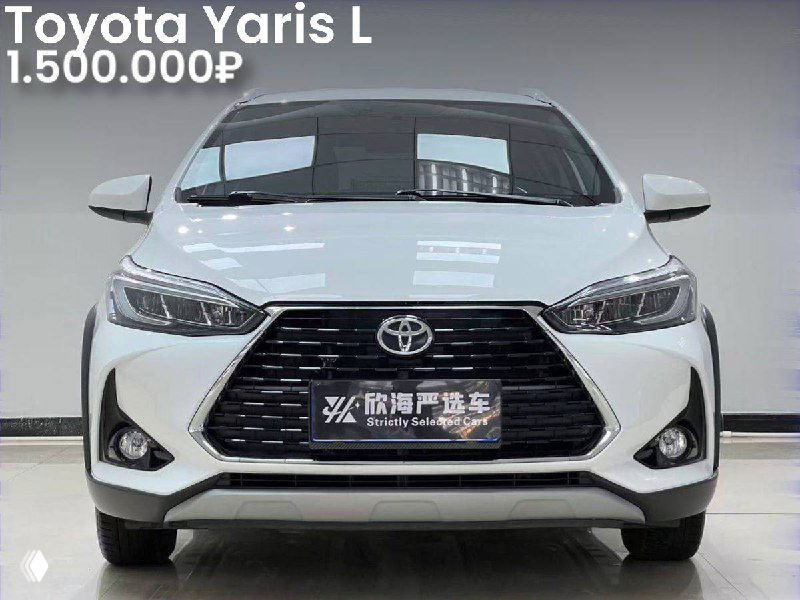Фронтальный снимок Toyota Yaris L белого цвета в автосалоне, видна решётка радиатора, фары и номерная табличка дилера на фото.
