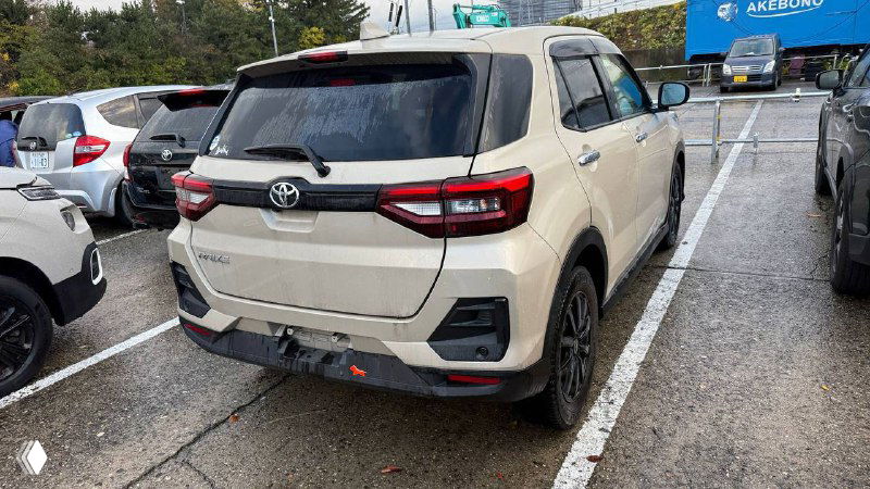 Задняя часть бежевого Toyota Raize 2021 на стоянке: багажник, задние фонари и общий вид автомобиля между соседними машинами на площадке.