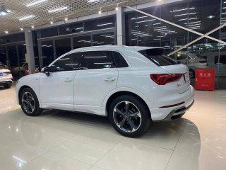 Белый Audi Q3 в выставочном зале, вид сзади-слева, видно форму задней части и колёсные диски на светлом фоне.