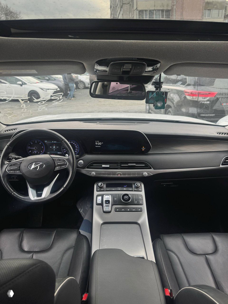 Вид салона Hyundai Palisade сверху: рулевое колесо, приборная панель и широкая центральная дисплейная панель в кабине с панорамной крышей на фоне улицы.