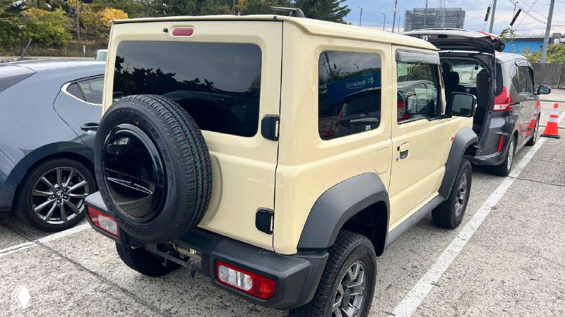Бежевый Suzuki Jimny Sierra 2021 — вид сзади: запасное колесо на двери, задние фонари, боковые окна и соседние автомобили на парковке.