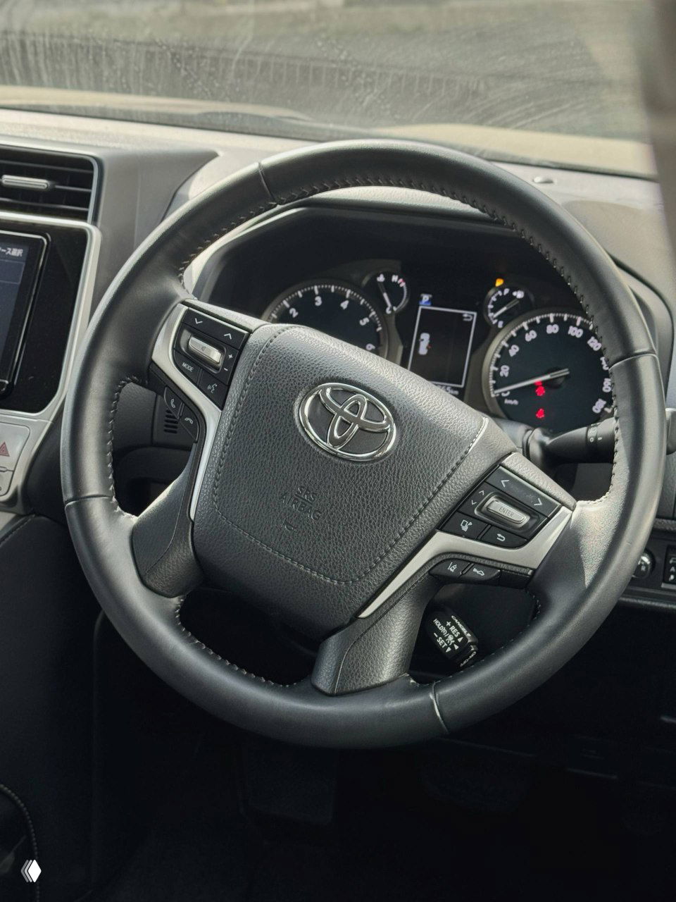 Крупный план рулевого колеса Toyota Land Cruiser Prado: вид логотипа, кнопки управления мультимедиа и часть приборной панели, салон в хорошем состоянии.