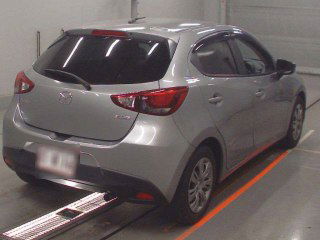 Серебристая Mazda Demio 2016 года, вид сзади в аукционном помещении Японии; видны фонари, багажник и задний бампер.