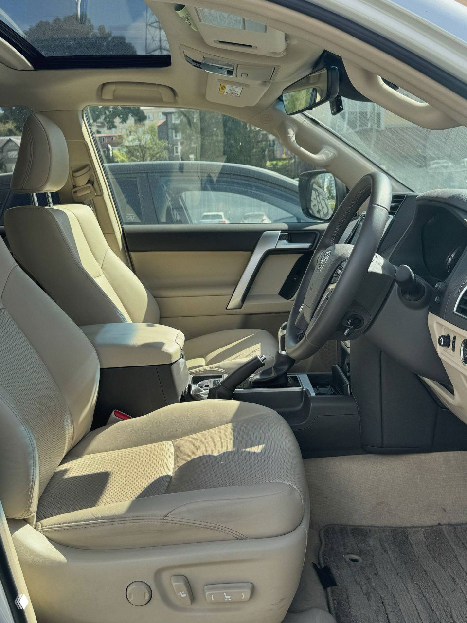 Передние сиденья и тоннель в салоне Prado 2021: светлая кожаная обивка, подлокотник и панорамная крыша создают представление о состоянии интерьера