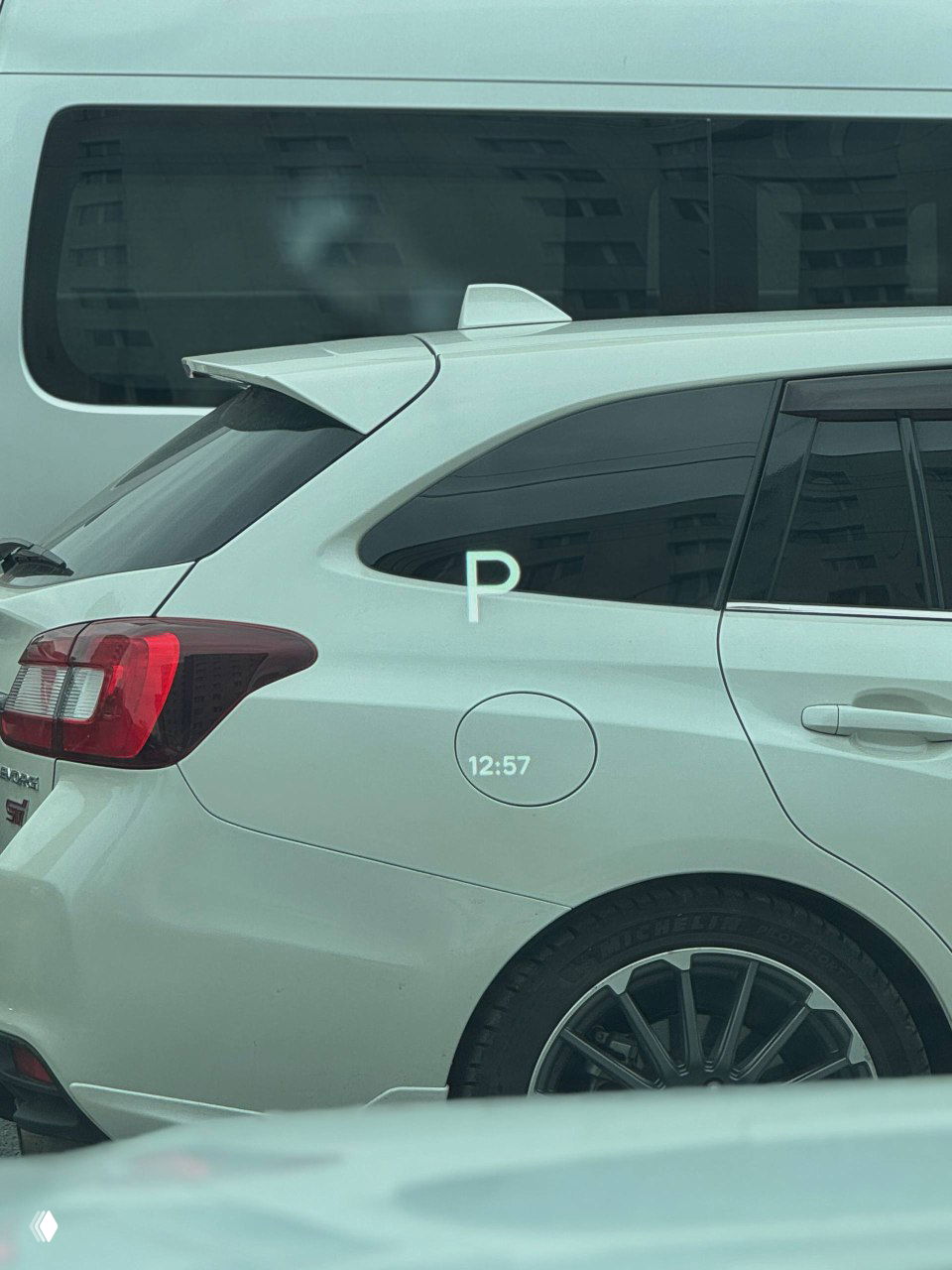 Задняя часть белого Lixiang L6, вид боковой двери и багажной зоны, наклейка про парковку и часть диска колеса на фоне городской застройки