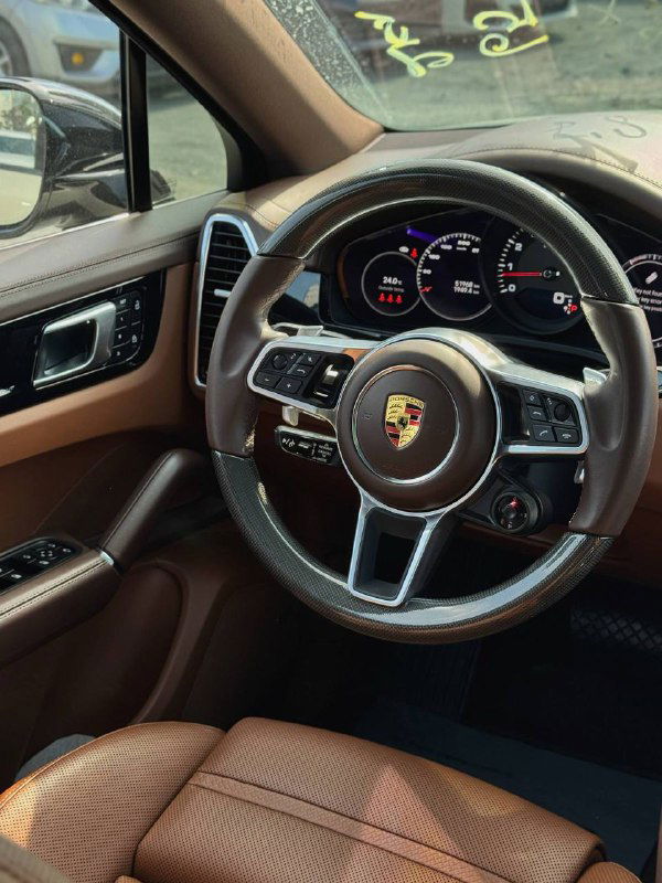 Крупный план руля Porsche с логотипом на натуральной коже и современной приборной панелью: вид центральной части салона и мультифункций на руле.