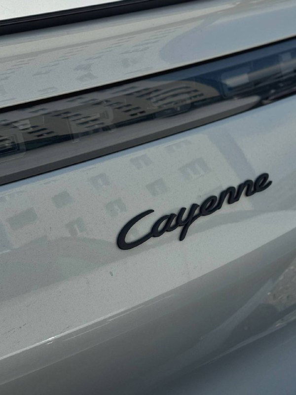 Эмблема «Cayenne» на задней двери автомобиля Porsche, крупный план шильдика и лакированной поверхности багажника с отражением зданий.