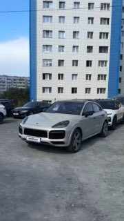 Porsche Cayenne светлого цвета, припаркованный на асфальтовой стоянке перед многоэтажным жилым домом; передний уголочный ракурс автомобиля в городском окружении.