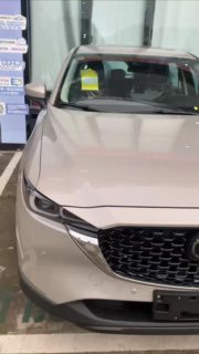 Частичный вид передней части золотистой/бежевой Mazda CX-5 на стоянке, видна фара, зеркало и наклейки на лобовом стекле