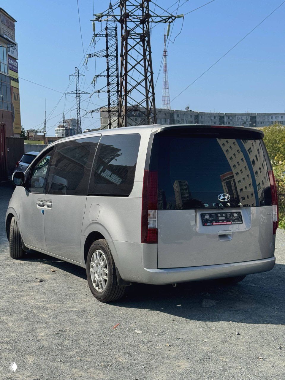 Hyundai Staria серебристого цвета вид сзади и под углом, видно крупные фонари, багажную дверь и логотип Hyundai на фоновой городской застройке