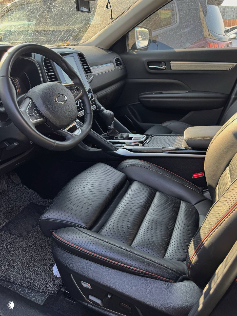 Вид салона Samsung QM6 с передними сиденьями, центральным тоннелем и рулём, чёрная кожаная обивка и обстановка дневной стоянки видна через стекло