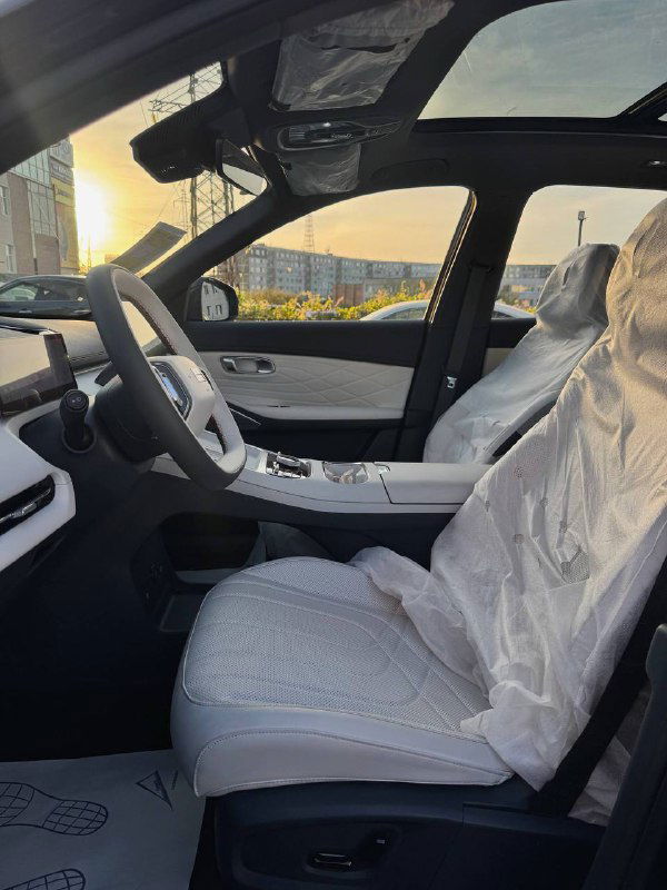 Передние сиденья Geely Galaxy Starship 7 в защитных чехлах, салон виден через открытое окно, на заднем плане городская застройка при тёплом свете