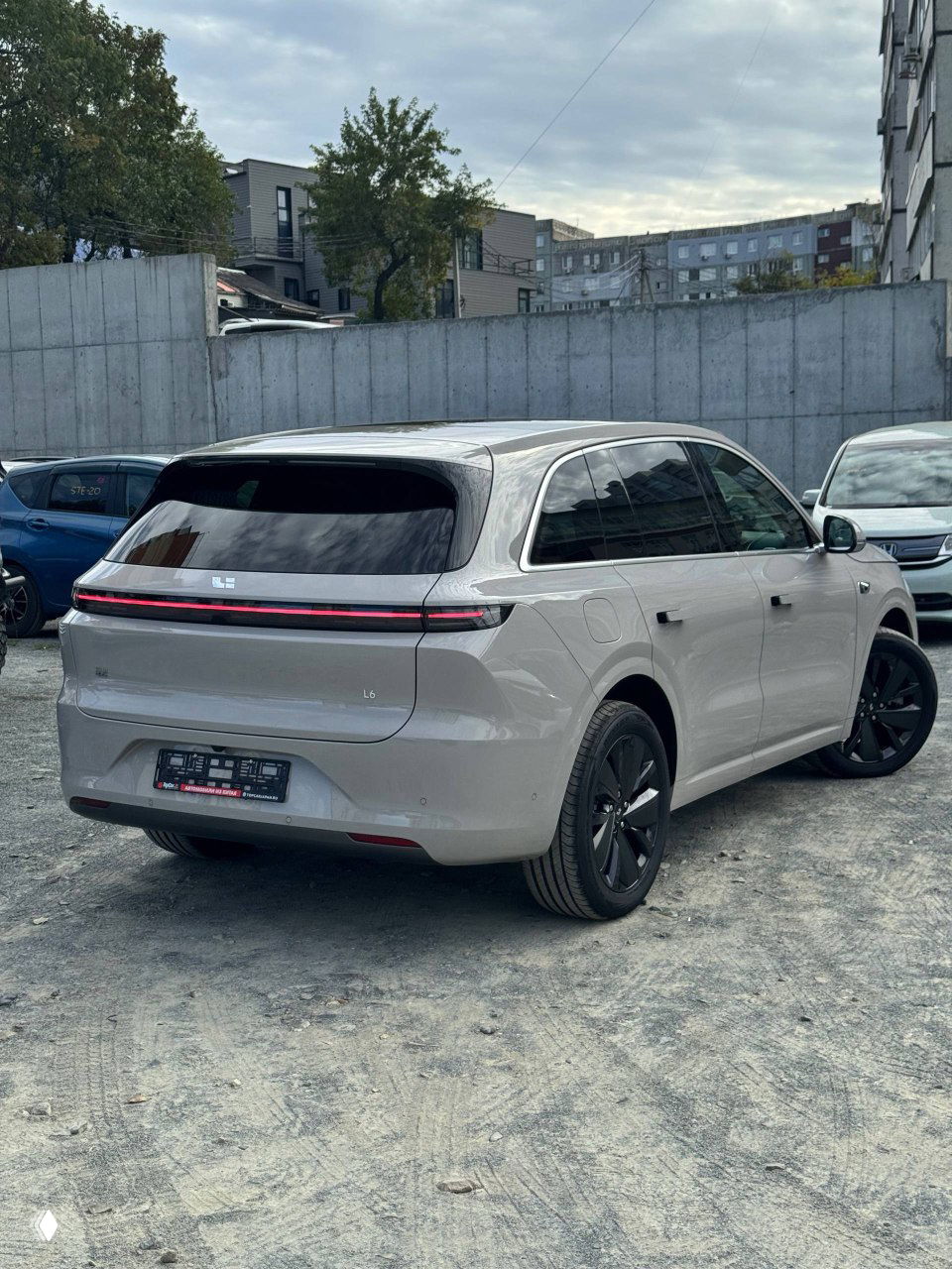 Lixiang L6 — задняя часть и боковой профиль автомобиля в слоновой кости, тонкая светодиодная световая полоса на багажнике, парковка на открытом воздухе с бетонным забором на заднем плане.