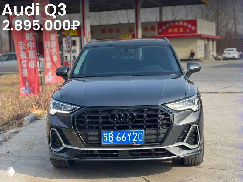 Тёмный Audi Q3 на открытой площадке дилера с китайскими плакатами на фоне; видно лицевую часть и номерную табличку, цена 2.895.000₽.