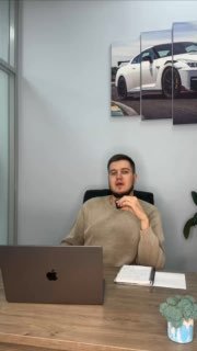 Мужчина за рабочим столом с ноутбуком в офисе, на стене крупный постер с легковым автомобилем — контекст видеообзора по рынку корейских авто.