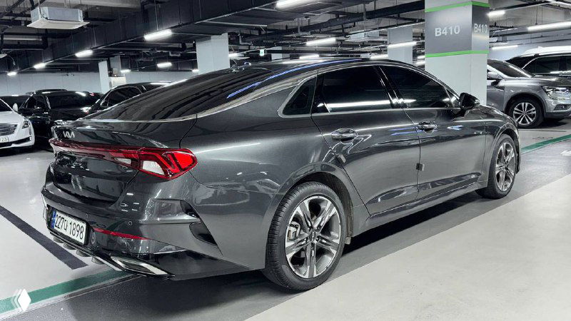 Боковой и задний ракурс KIA K5 2023 в крытой стоянке: вид сзади‑сбоку, окраска тёмно‑серого металлика и задние фонари автомобиля