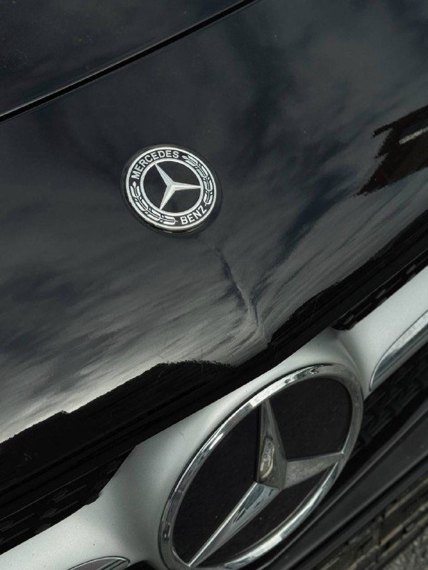 Крупный план эмблемы Mercedes на капоте чёрного автомобиля, отражение неба на лаковой поверхности кузова и вид центрального логотипа.