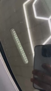 Крупный план VIN на лобовом стекле Volkswagen Golf с отражением геометрического потолочного освещения и тенью руки фотографа — видимость идентификационного номера.