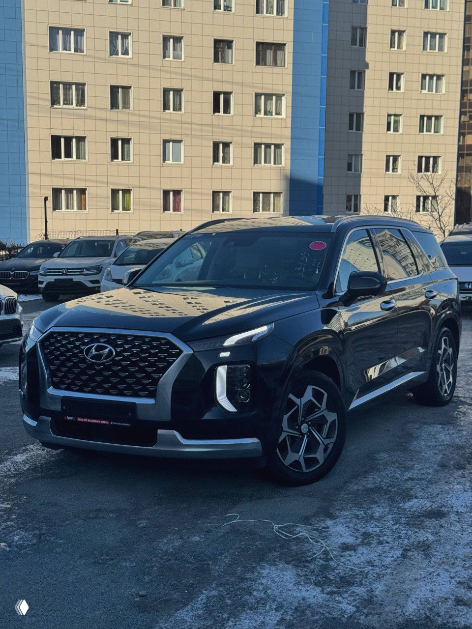 Передняя часть чёрного Hyundai Palisade, вид спереди на парковке у жилого дома; видны фары, решётка радиатора и зимняя дорога с наледью.