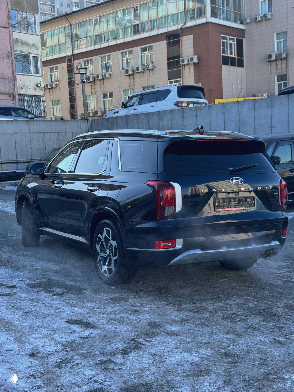 Задняя часть чёрного Hyundai Palisade на открытой парковке с легким паром от выхлопа; видна современная задняя оптика и номерной контейнер.