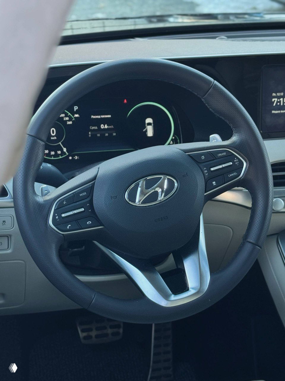 Крупный план рулевого колеса Hyundai с логотипом марки, элементы управления на руле и часть цифровой приборной панели на фоне салона.