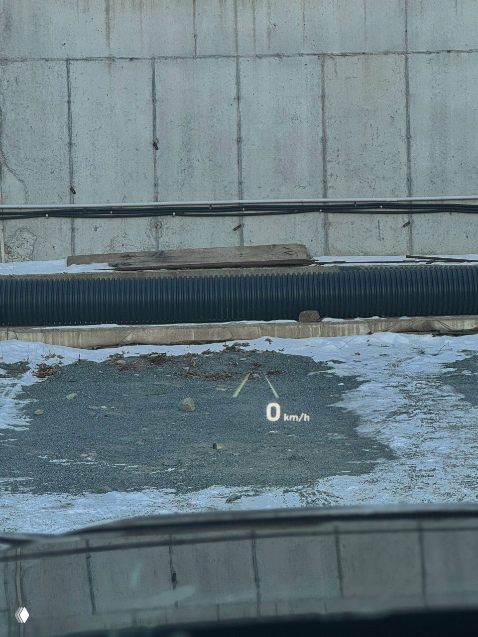 Проекция скорости (HUD) на лобовом стекле автомобиля, видимая на фоне снежного покрытия и бетонной стенки на стоянке.