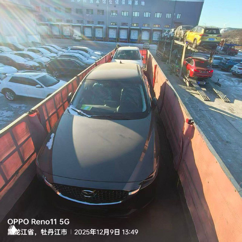 Вид сверху на тёмную Mazda CX-5, закреплённую на платформе автовоза; вокруг другие автомобили и солнечный блик на кузове в морозный день.