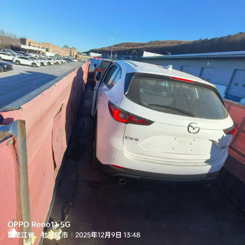 Фото сзади-боковой части белой Mazda CX-5 на автовозе; видна дорожная поверхность, ограждения платформы и общий контекст погрузки.