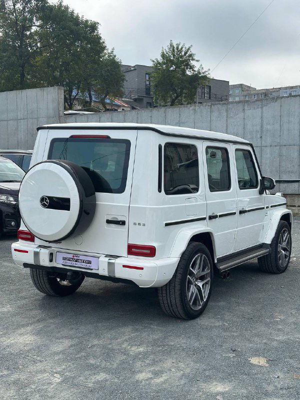 Задний вид белого Mercedes G63 AMG на стоянке, видна запаска на двери багажника, задние фонари и общий городской фон автомобиля.
