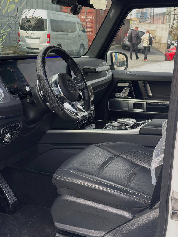 Ещё один ракурс передних кресел и рулевой колонки Mercedes G63 AMG, видна чёрная кожа, декоративные строчки и педали внизу.