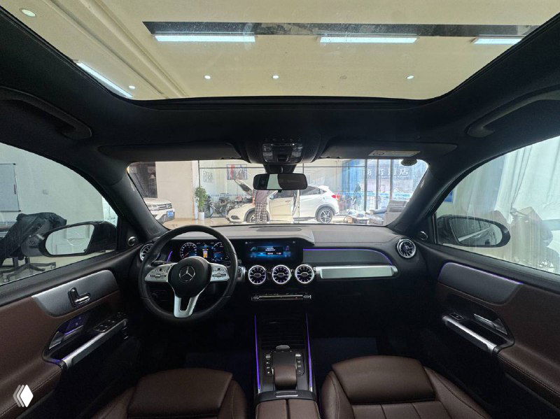 Вид салона Mercedes GLB через лобовое стекло: панорамная крыша, приборная панель и хороший обзор торпедо в дневном освещении шоурума.