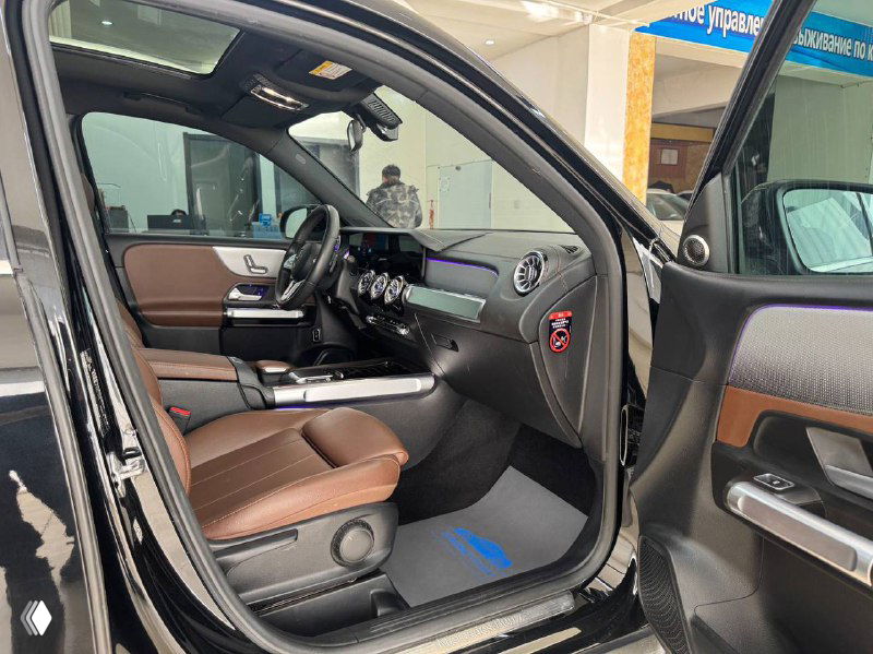Открытая дверь водителя и вид переднего ряда сидений Mercedes GLB: коричневая кожаная отделка, порожек и панель управления на двери видны крупным планом.