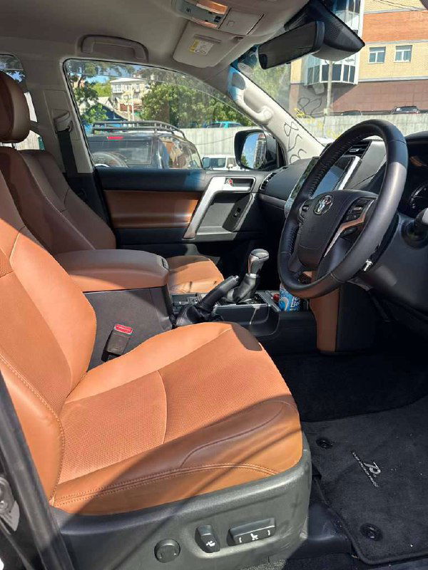 Передние сиденья Toyota Prado в коричневой кожаной обивке — крупный план водительского сиденья и часть передней панели салона с подлокотником.