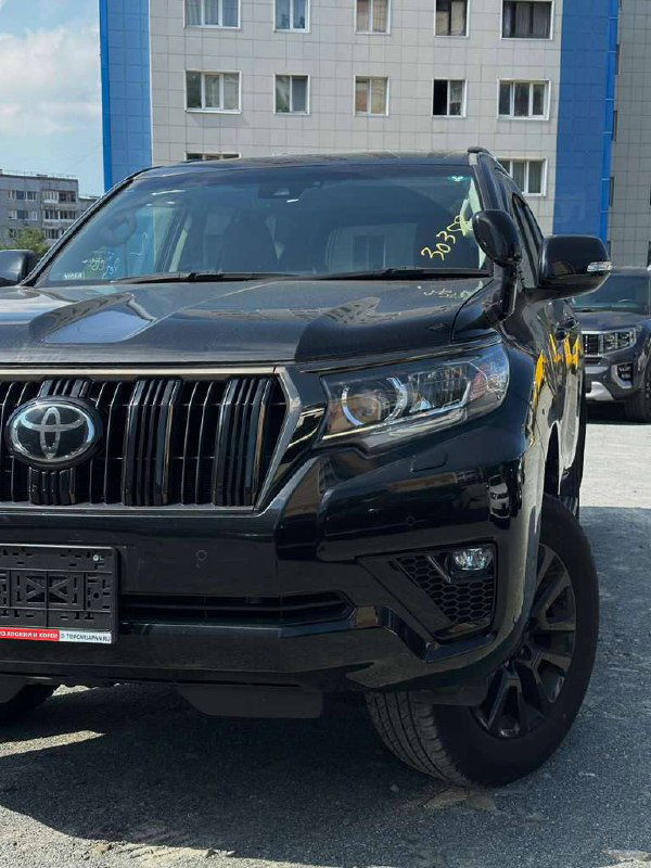Фронтальный вид на переднюю часть Toyota Prado с акцентом на фары и капот — чёрный внедорожник стоит на площадке возле зданий.