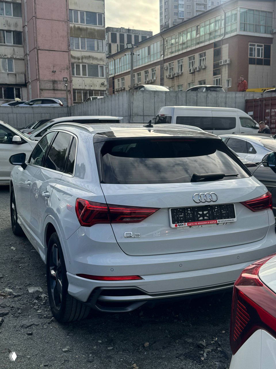 Тыловая часть белого Audi Q3 на стоянке; видно багажник, эмблему Audi и модельный шильдик Q3, фонари и номерную рамку.