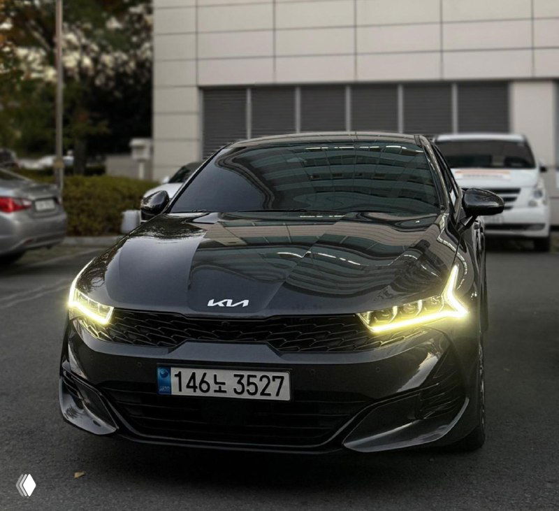 Фронтальный крупный план чёрного KIA K5, акцент на фирменной решётке и угловых светодиодных фарах, автомобиль на городской парковке с отражениями на капоте.