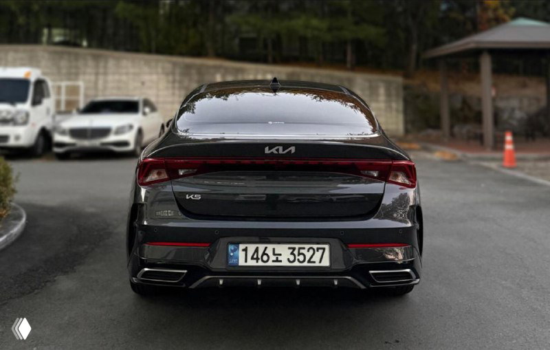 Вид сзади на чёрный KIA K5: кузов, задние фонари и номер тормозной парковки, машина стоит на асфальтовой площадке с городской оградой на заднем плане.