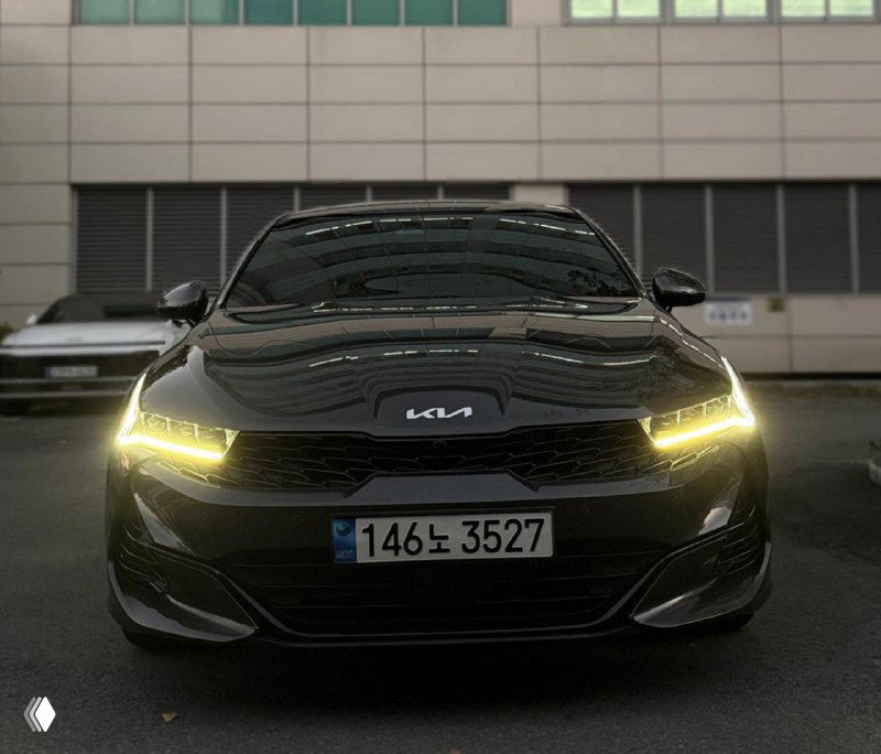 Ещё один фронтальный вид KIA K5 в тёмном исполнении с подчёркнутыми дневными ходовыми огнями на фоне офисного фасада, подчёркнут спортивный стиль автомобиля.