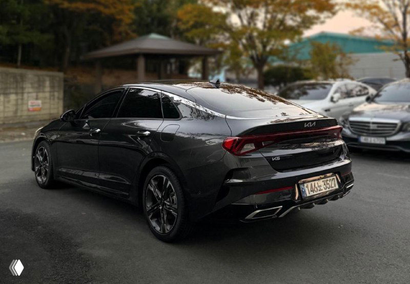 Ещё один снимок сзади и сбоку чёрного KIA K5: подчёркнута линия крыши и кормовая часть, автомобиль находится на стоянке среди других машин под деревьями.