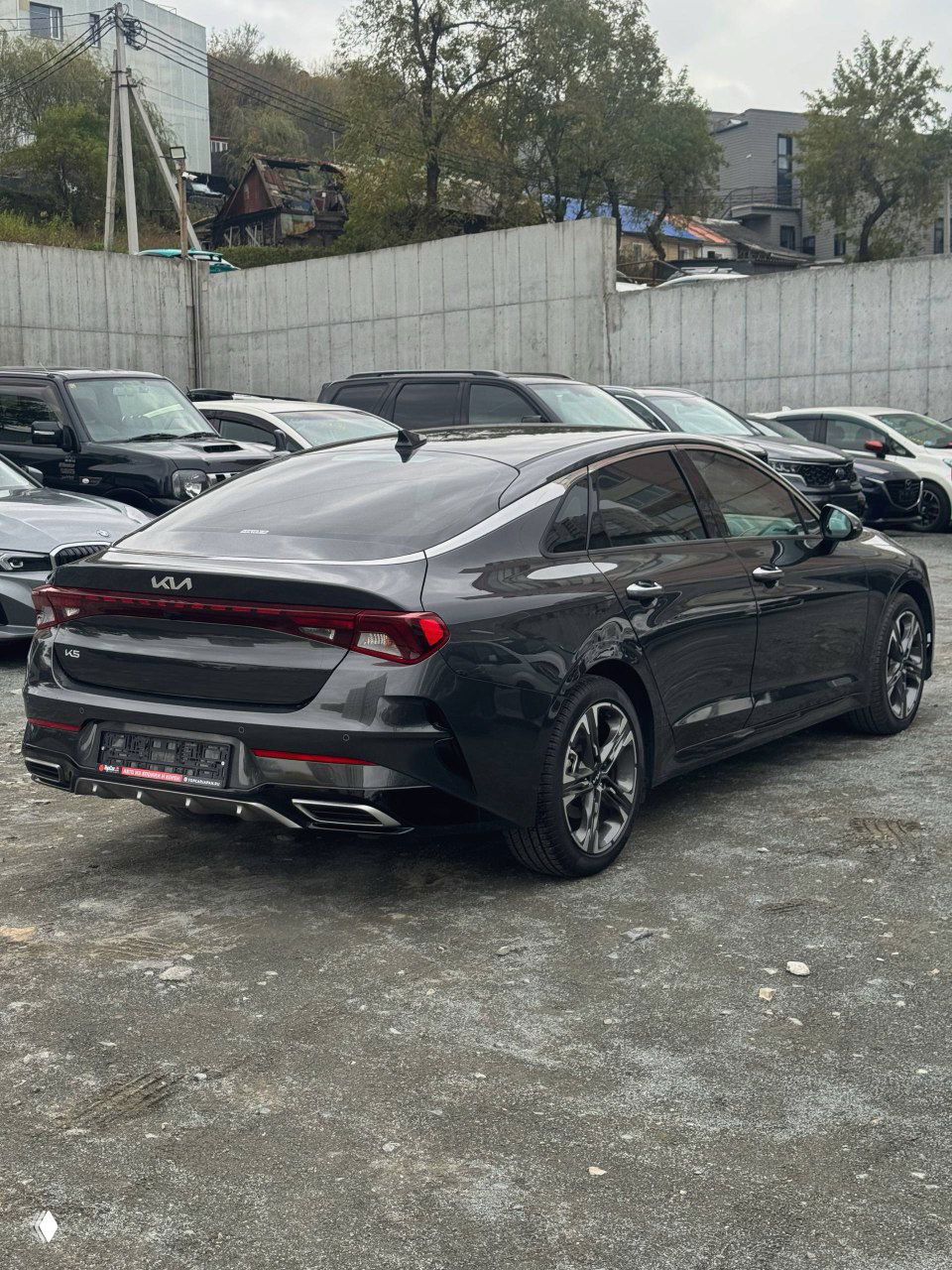 Задняя часть KIA K5 2023 с диодной полосой стоп-сигналов и спортивным бампером, общий вид автомобиля на стоянке.