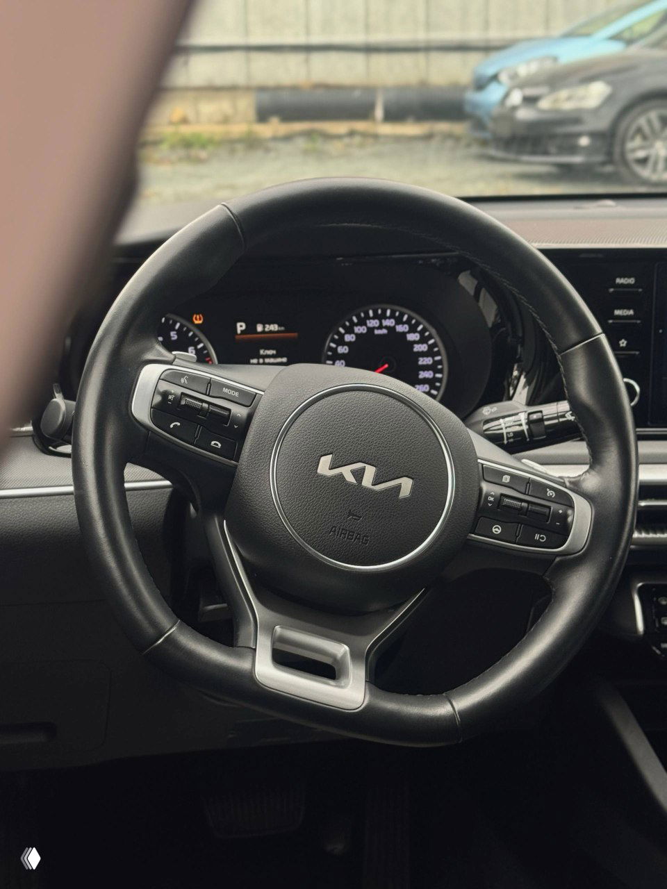 Рулевое колесо KIA с новым логотипом, кнопки управления мультимедиа и приборная панель видны на фоне лобового стекла.